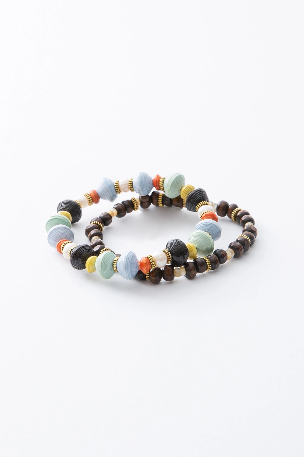 The two bracelets included in the Ubuntu set are shown stacked. Both are stretchy, single-strand beaded bracelets. One bracelet is strung with hand rolled paper beads in shades of light blue, mint, orange, yellow, and black, along with decorative brass beads. The other bracelet is strung with dark brown ox horn beads and fresh water pearl beads, accented with the same decorative brass beads.