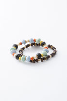 The two bracelets included in the Ubuntu set are shown stacked. Both are stretchy, single-strand beaded bracelets. One bracelet is strung with hand rolled paper beads in shades of light blue, mint, orange, yellow, and black, along with decorative brass beads. The other bracelet is strung with dark brown ox horn beads and fresh water pearl beads, accented with the same decorative brass beads.