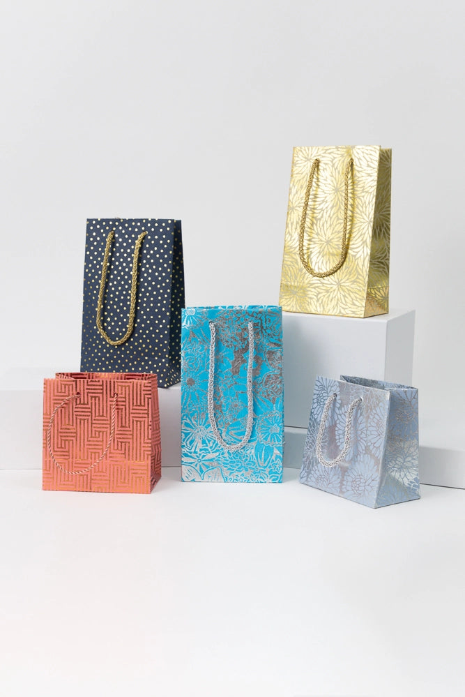 Five examples of the Upcycled Metallic Gift Bags are pictured. Some are tall and rectangular, others are short and square. Each has two woven cord handles in a complementary color attached to the top of the bag. The bags are each a different color and pattern, but all have metallic elements that give the paper a shiny look.