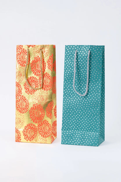 Two examples of the Upcycled Metallic Wine Bags are pictured. Both are tall, skinny, and rectangular. Each has two woven cord handles in a complementary color attached to the top of the bag. The bags are each a different color and pattern, but both have metallic elements that give the paper a shiny look.