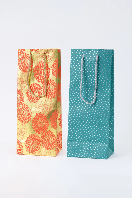 Two examples of the Upcycled Metallic Wine Bags are pictured. Both are tall, skinny, and rectangular. Each has two woven cord handles in a complementary color attached to the top of the bag. The bags are each a different color and pattern, but both have metallic elements that give the paper a shiny look.