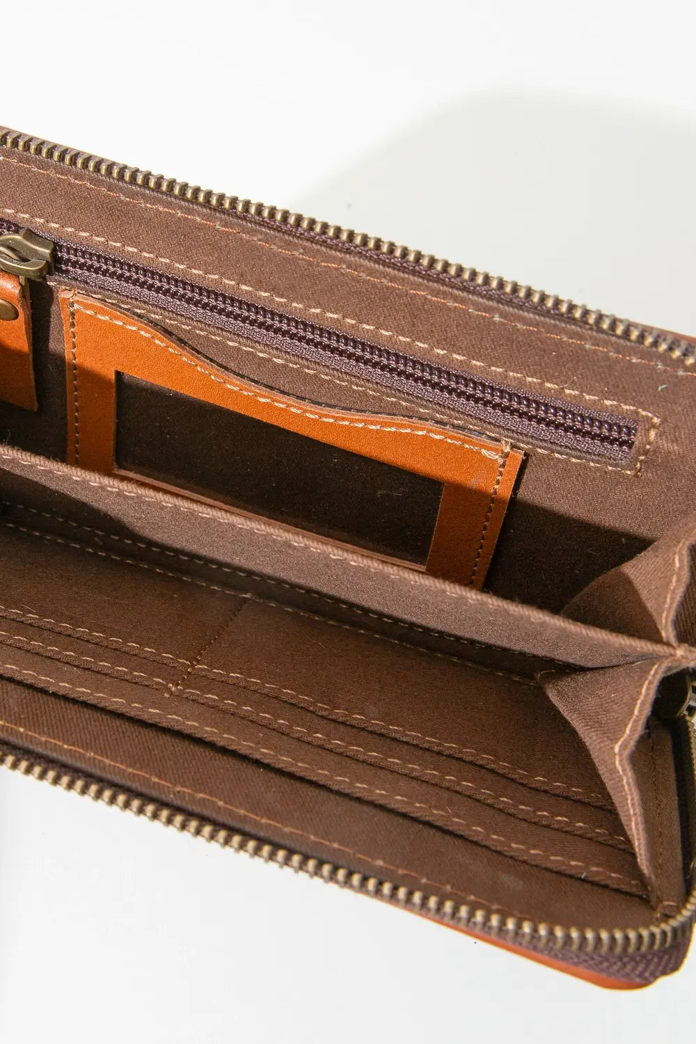 The Vaquera Wallet is shown unzipped. One interior wall has a zip pocket, as well as an ID slot pocket that is clear in the center and outlined in orange leather. There is a divider in the center of the wallet, dividing it into two main compartments. The opposite wall has several fabric slot pockets for cards.