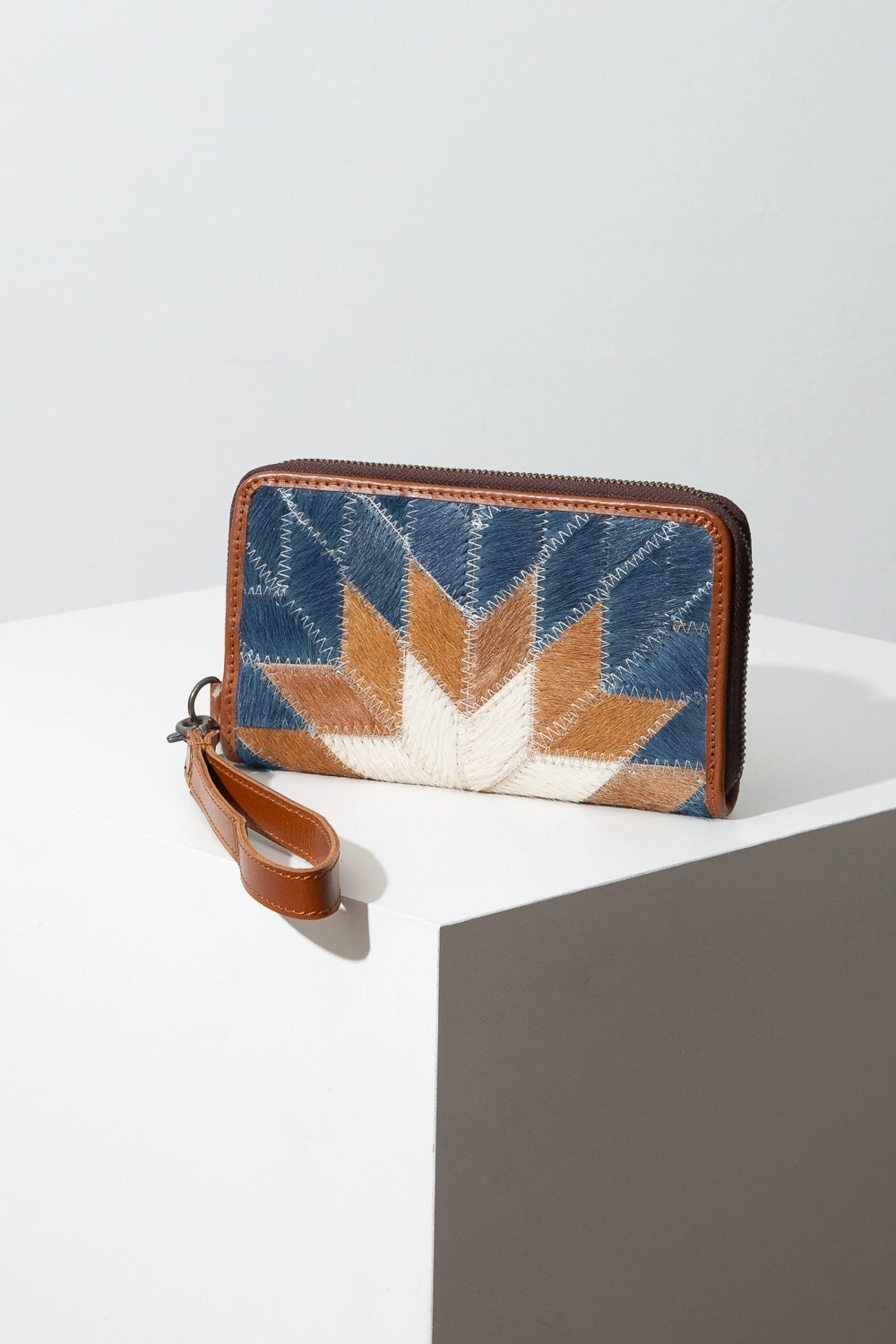The Vaquera Wallet is a rectangular leather wallet that is wider than it is tall. A zipper runs along the left side, top, and right side of the wallet. There is a brown leather wrist strap clipped to the zipper pull that can be removed. The wallet is composed of diamonds of white, brown, and blue leather stitched together to form a quilt-style starburst pattern.