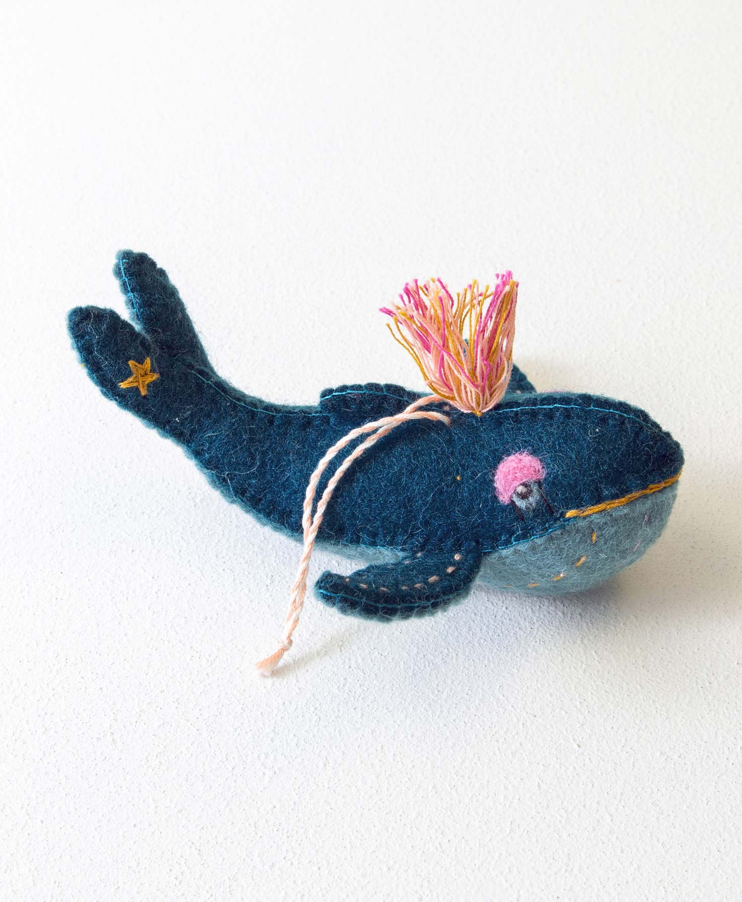 The Wonder Whale Ornament is a three-dimensional whale composed of wool. The top half of the whale is dark blue and the bottom half is light blue. Handstitched details run along the sides of the whale. It has felt eyes and has a bright tassel on top of its head. The ornament is finished with a twisted cord loop where it can be hung.