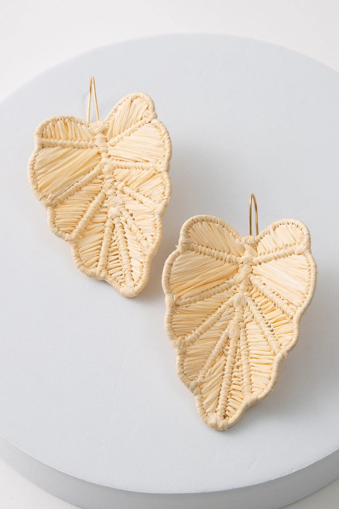 The Woven Palm Earrings are large statement earrings composed of natural, undyed raffia woven into a tropical leaf shape. The leaves are attached to gold ear hooks.